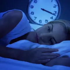 Kobieta leży w łóżku, patrząc w sufit. Zegar obok pokazuje późną noc. To obraz tego, jak brak snu wpływa na mózg.
