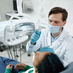 Dentysta w masce i rękawiczkach bada pacjenta w gabinecie stomatologicznym.