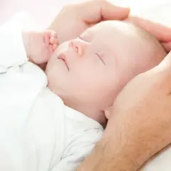 Dłoń ojca delikatnie obejmuje główkę śpiącego niemowlęcia. Dziecko, mimo że ma niską masę urodzeniową, wygląda na spokojne i bezpieczne.
