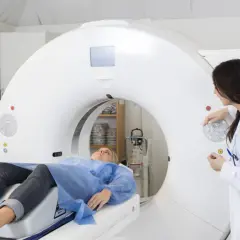 Pacjentka leży na stole tomografu, a lekarka obsługuje konsolę. Badanie ma na celu wykrycie ewentualnych dolegliwości po podaniu kontrastu.