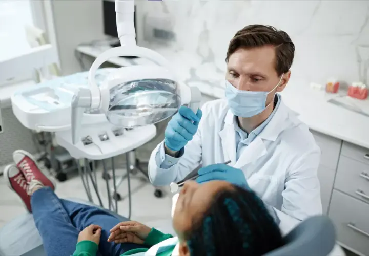 Dentysta w masce i rękawiczkach bada pacjenta w gabinecie stomatologicznym.