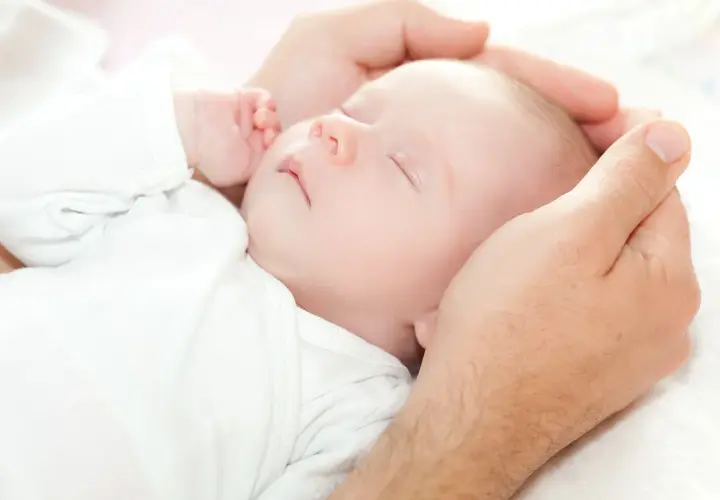 Dłoń ojca delikatnie obejmuje główkę śpiącego niemowlęcia. Dziecko, mimo że ma niską masę urodzeniową, wygląda na spokojne i bezpieczne.