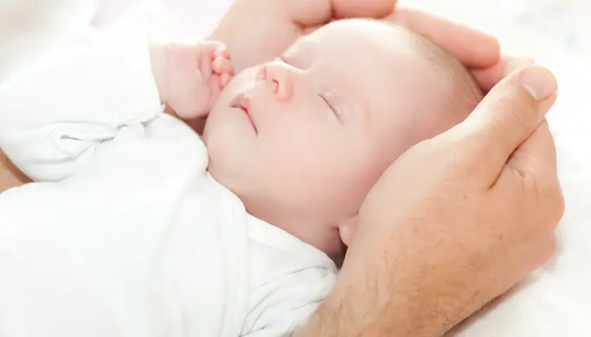 Dłoń ojca delikatnie obejmuje główkę śpiącego niemowlęcia. Dziecko, mimo że ma niską masę urodzeniową, wygląda na spokojne i bezpieczne.