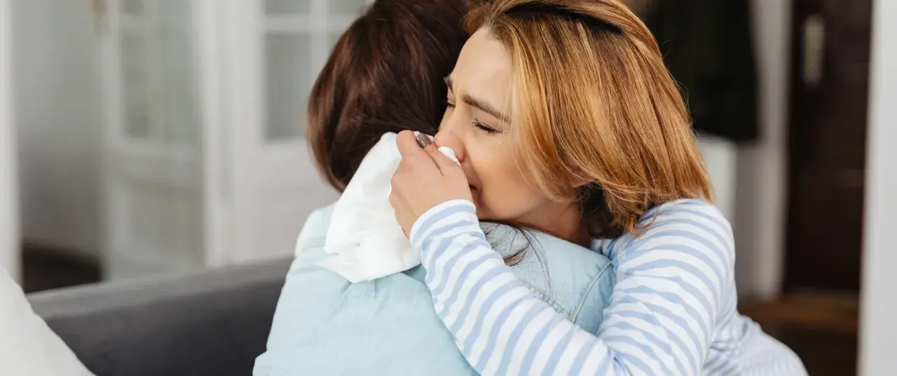 Kobieta pociesza inną, która płacze, wycierając nos chusteczką. Scena sugeruje lęk separacyjny u dorosłych i potrzebę wsparcia.