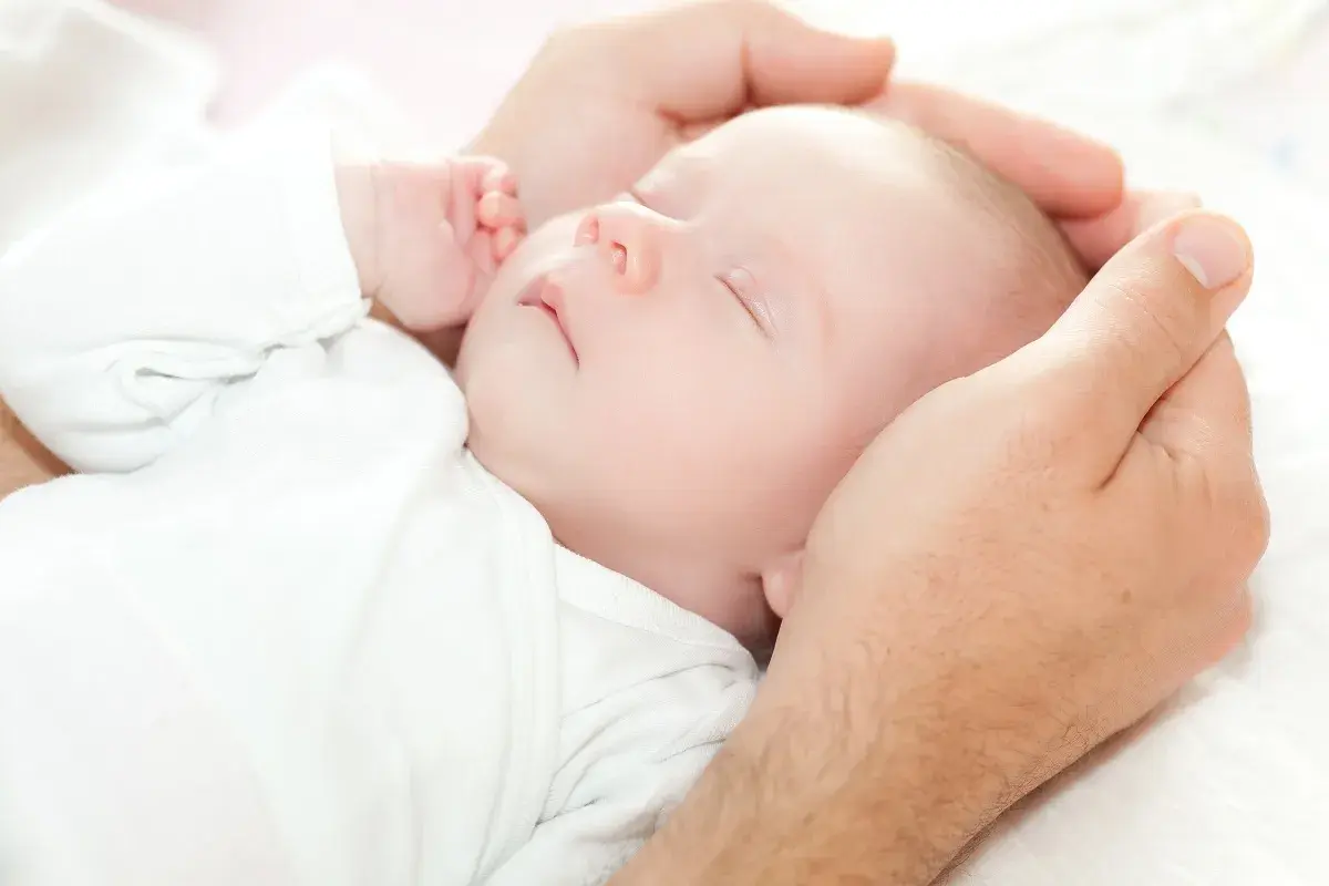 Dłoń ojca delikatnie obejmuje główkę śpiącego niemowlęcia. Dziecko, mimo że ma niską masę urodzeniową, wygląda na spokojne i bezpieczne.