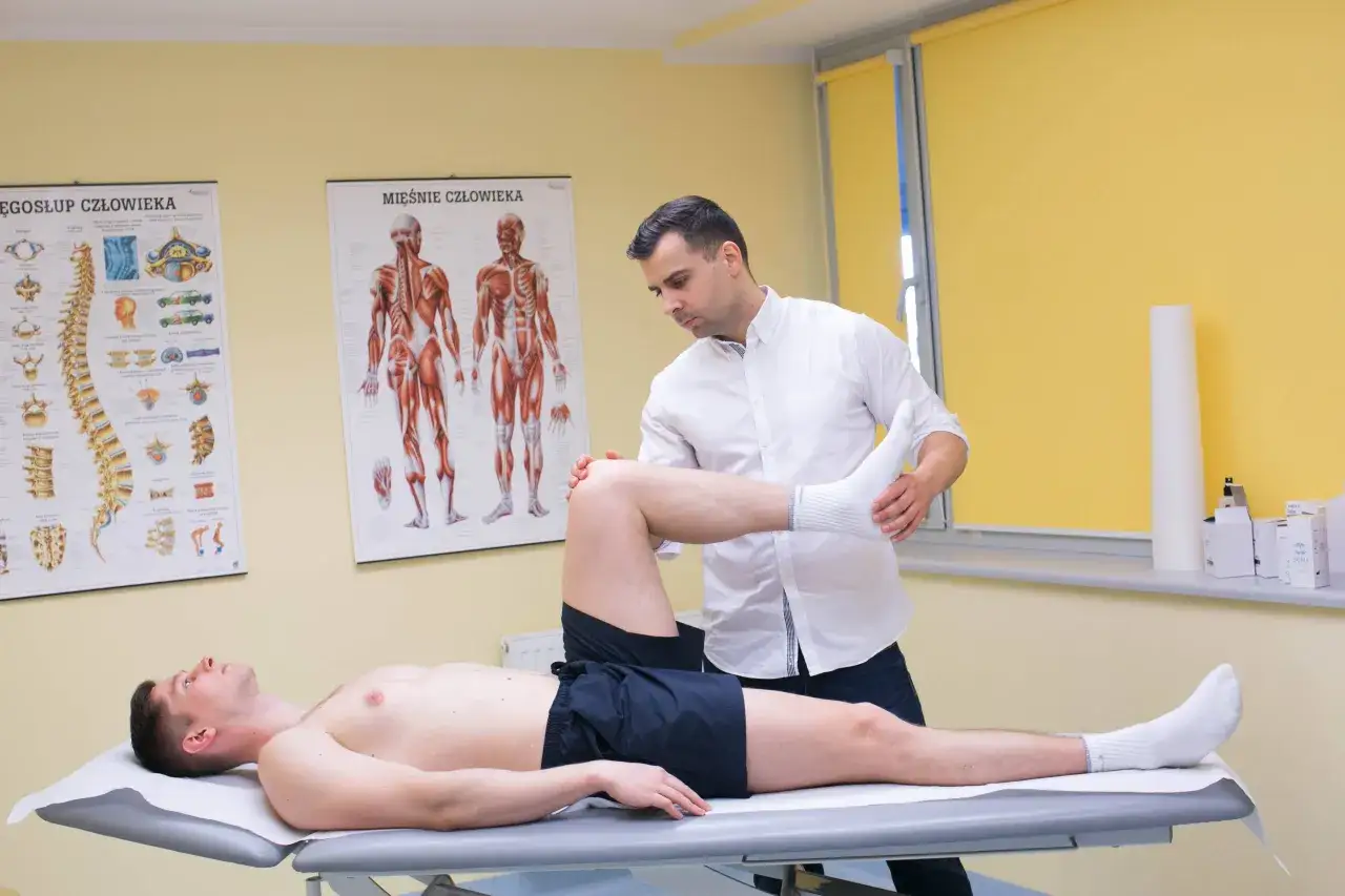 Fizjoterapeuta bada nogę pacjenta, szukając rozwiązania co na naderwany mięsień. Na ścianie wiszą anatomiczne plakaty.