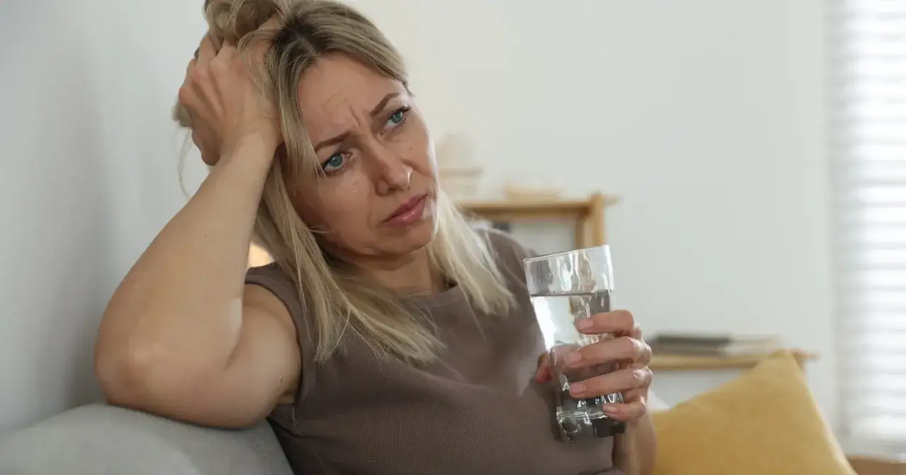 Kobieta z ręką na głowie, zmartwiona, trzyma szklankę wody. Może to być jeden z objawów okresu przedmenopauzalnego.